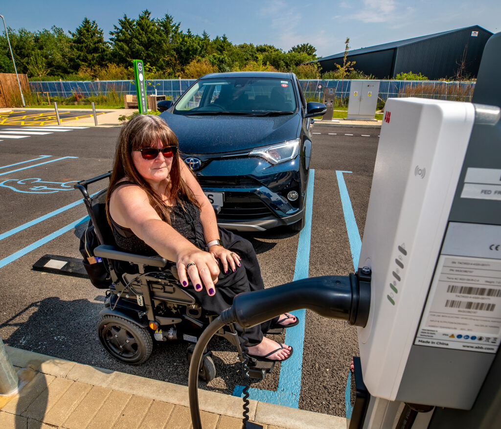 Photo of a woman sitting in a powered wheelchair and reaching towards the connector of an EV charging cable plugged into a charging unit mounted on a kerb. The kerb prevents her from getting close enough to touch the connector.