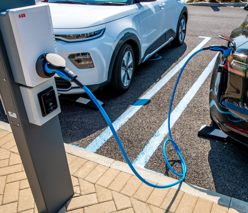 Photo showing a cable connecting a socket on the rear wing of a car to a socket on a charging unit mounted on a post in a car park.