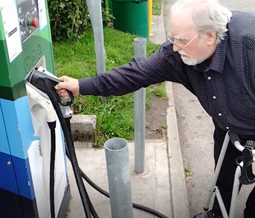 Photo of an older man standing facing reaching forwards to hold the charging connector that is holstered in the front of a rapid charging unit in an outdoor car park. The charging unit is set back on a raised kerb, so he is leaning forward over his walking frame, to reach the connector. He cannot get closer to the unit because his walking frame is at the edge of the kerb.