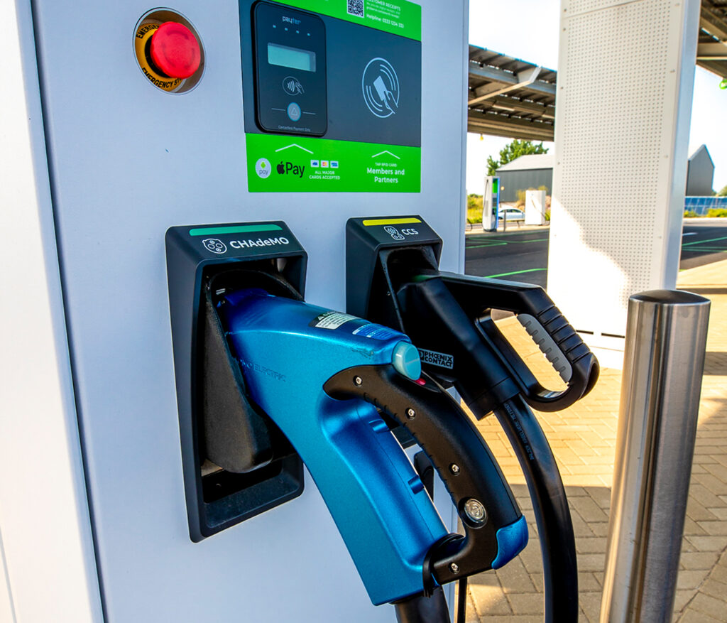 Photo showing a large white charging unit shaped like a modern petrol pump. Two different labelled connectors (CHAdeMO and CCS) with handles rest in the front face of the unit ready to be used, and a red emergency stop button and contactless payment features are also visible.