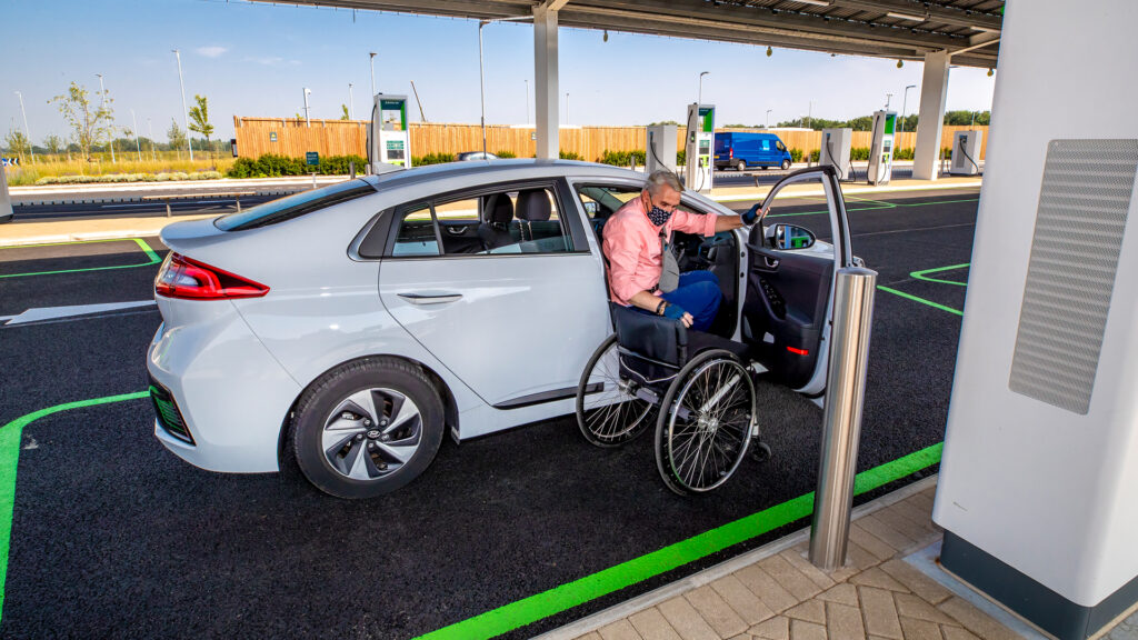 Photo of a man sitting in the driver’s seat of a car with the door open. He holds the edge of the door frame with one hand and uses the other to reach across to his manual wheelchair which sits just outside the car door, preparing to transfer from the driver’s seat into the wheelchair.