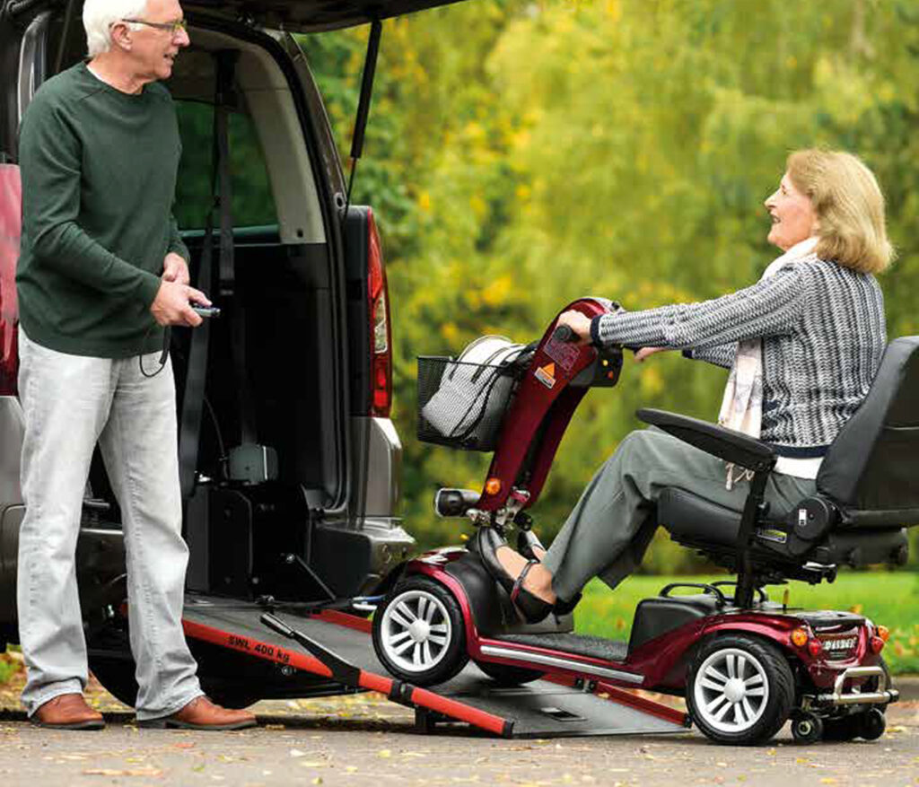 A woman drives her mobility scooter onto the ramp that sticks out from the back of a van that has its tailgate open. She looks towards a man who is standing next to the ramp.