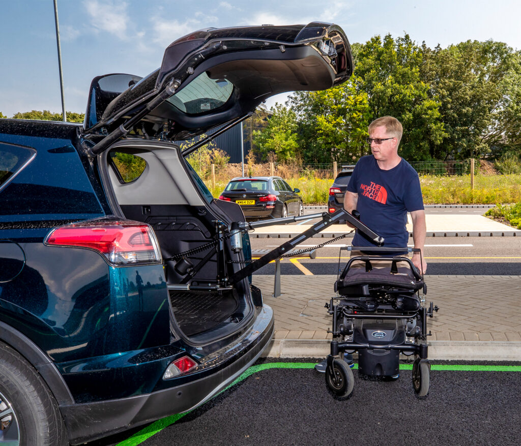 Photo of a man wearing sunglasses standing in car park behind a large car with its tailgate open. He uses a remote control to operate the boot hoist which is suspending a powered wheelchair out of the back of the boot on an arm attached to the inside of the boot.