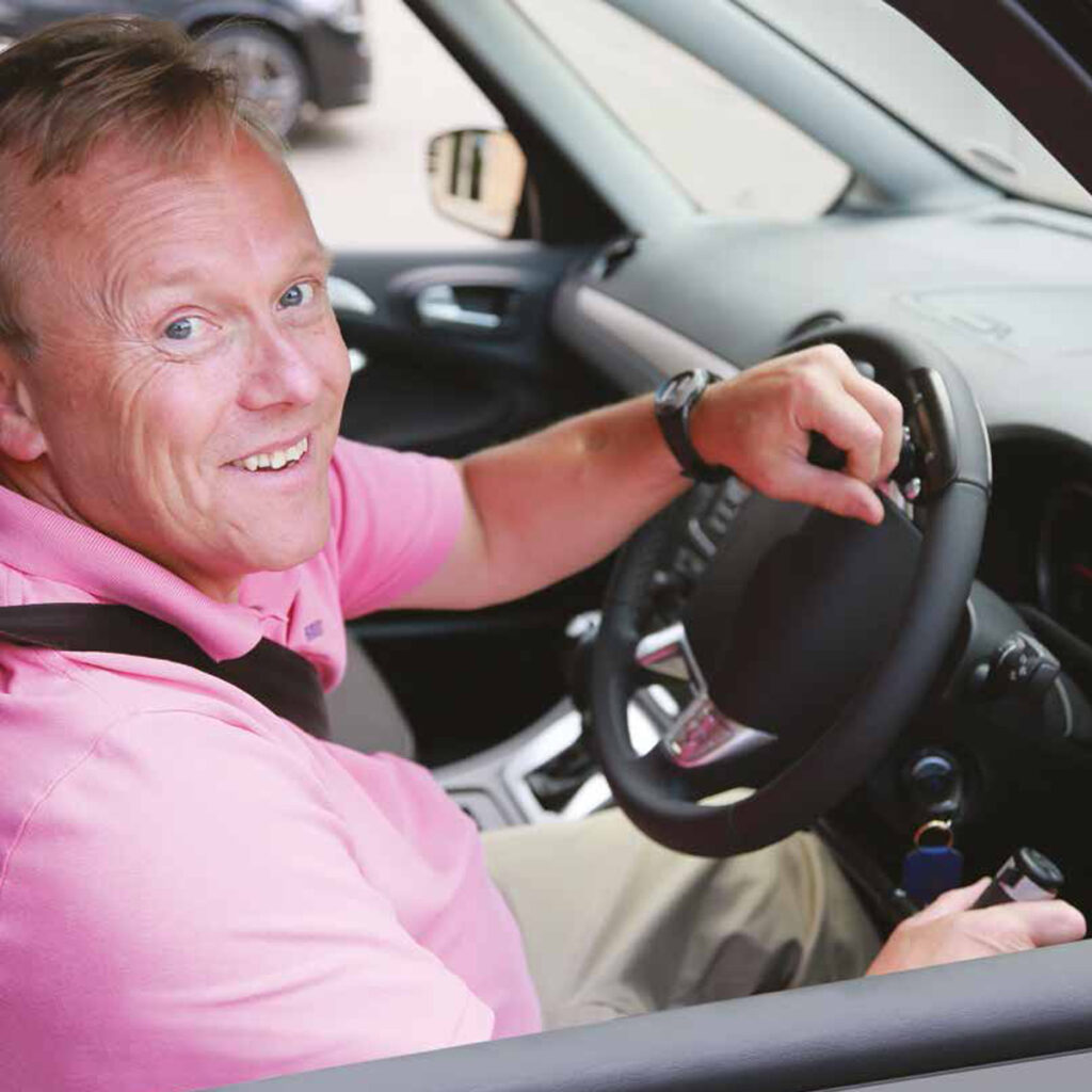 Photo of a man sitting in the driver’s seat of a car that has some hand controls. He is wearing a seatbelt and resting his hands on the hand controls as he smiles towards the camera.