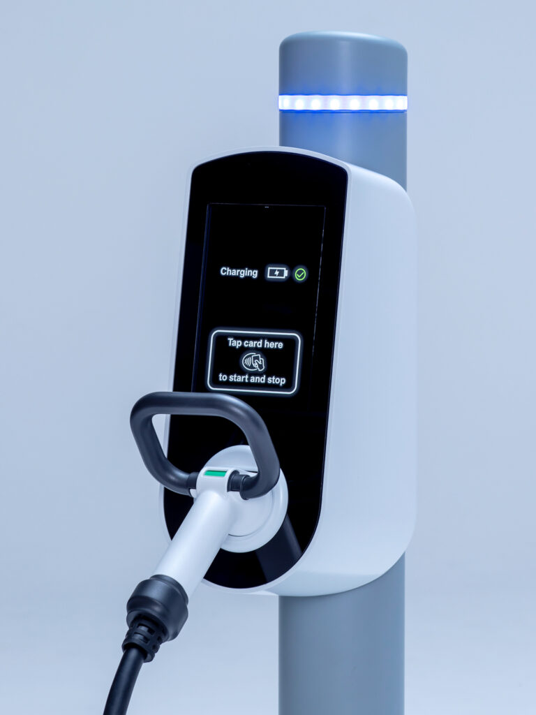 Close up photo of the front of a small EV charging unit. It is a curved white box with a tilted flat black front, mounted on a grey cylindrical post. There is an EV charging connector plugged into the white socket on at the bottom of the front face, that has a grey curved loop handle sticking up and out from the back of the connector. The black screen displays illuminated white text that says “charging” next to a white battery symbol and a green tick. There is also an illuminated white card-shaped rectangular outline that says “Tap card here to start and stop” with a white contactless symbol.