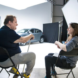 Photo of a man backstage at a photography studio sitting on a chair resting his hand on the top of a metal walking stick in front of him. He is having a conversation with one of Designability’s researchers who sits facing towards him.