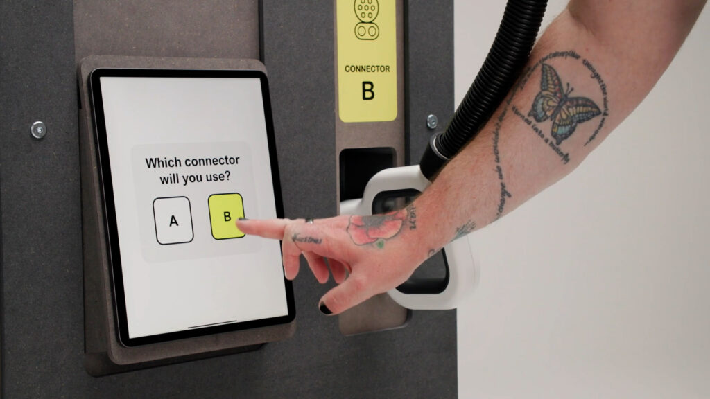 Close up photo of a hand reaching to touch the screen on the front of a rapid charging unit. The white screen asks, which connector will you use? and shows a white square labelled A in black text and a yellow square labelled B in black text. The connector being gripped is labelled connector B on a yellow label on the front of the charging unit.