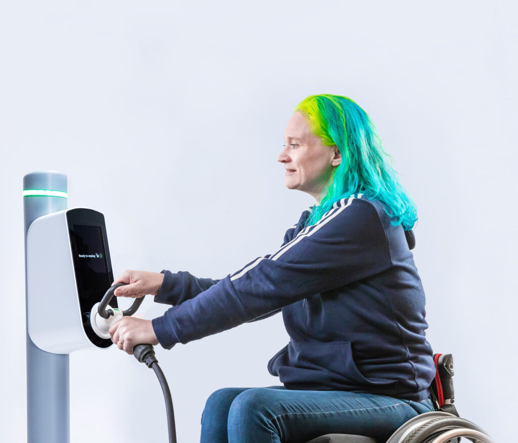 Photo of a woman sitting in a manual wheelchair in front of a simple EV charging unit that has a connector plugged into the socket on the front of it at waist height. She reaches forwards and holds the connector with two hands, by its main part and by the looped handle on the top of the connector.